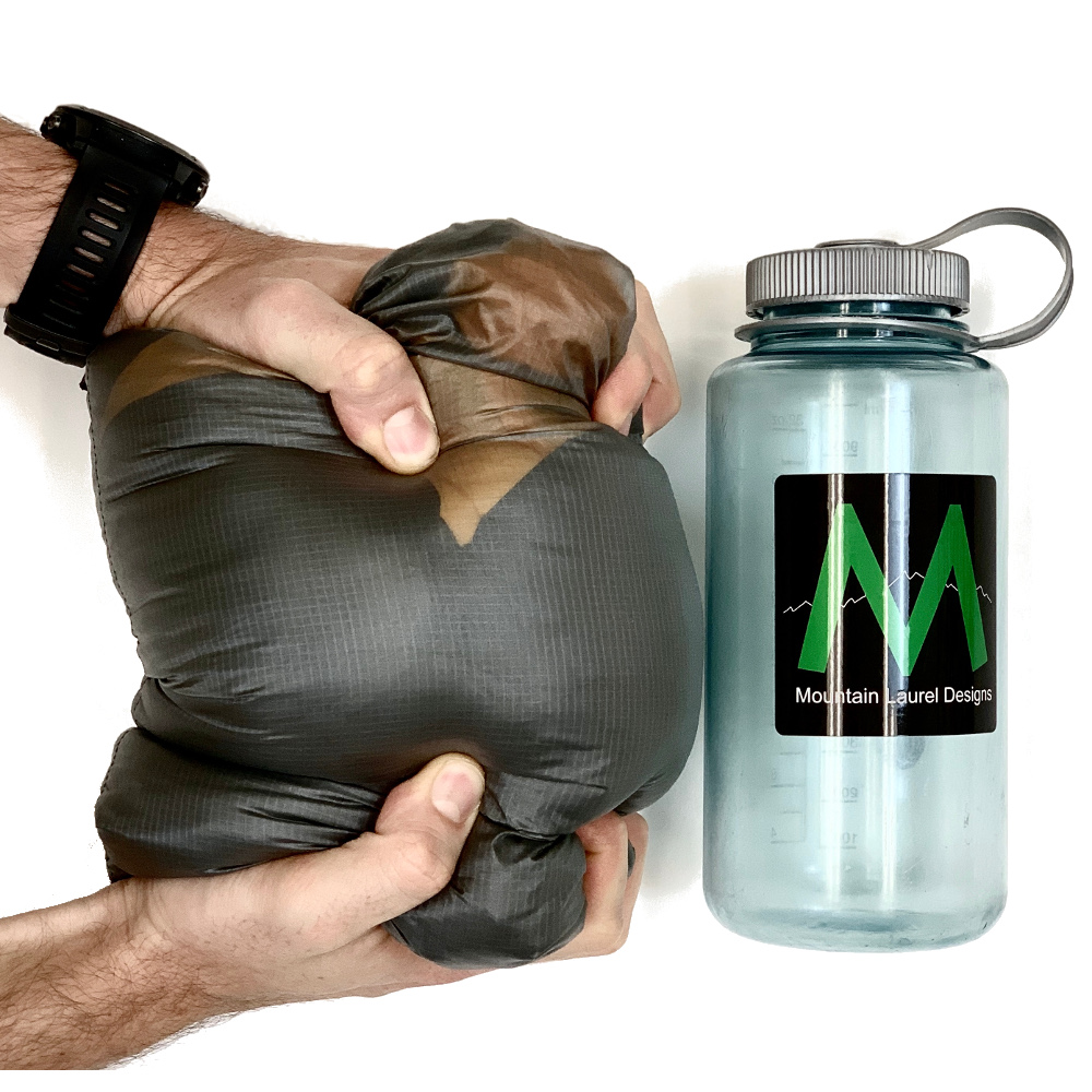 The Vision Quilt packed in its stuff sack next to a Nalgene bottle for a size comparison.A man is compressing it with his hands and it is roughly the same size as the Nalgene